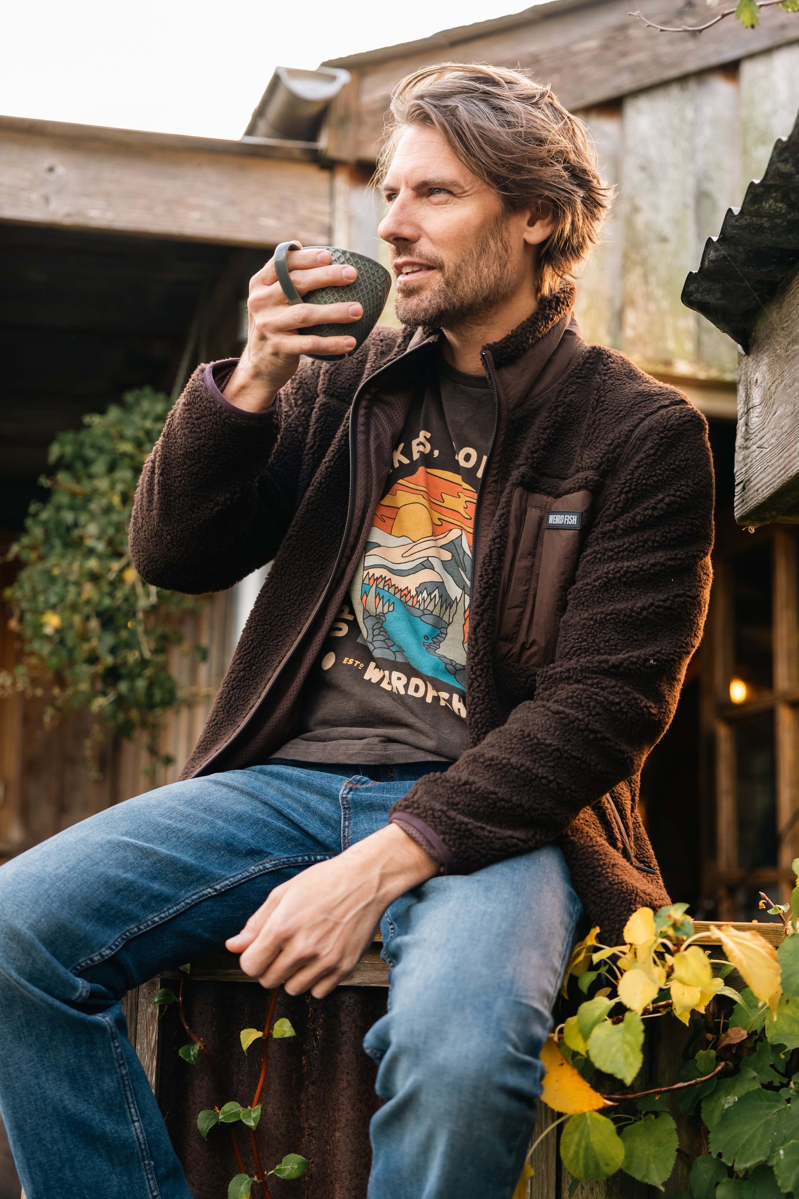 Man in the Weird Fish Fletcher full zip borg fleece in Chocolate sitting with a drink